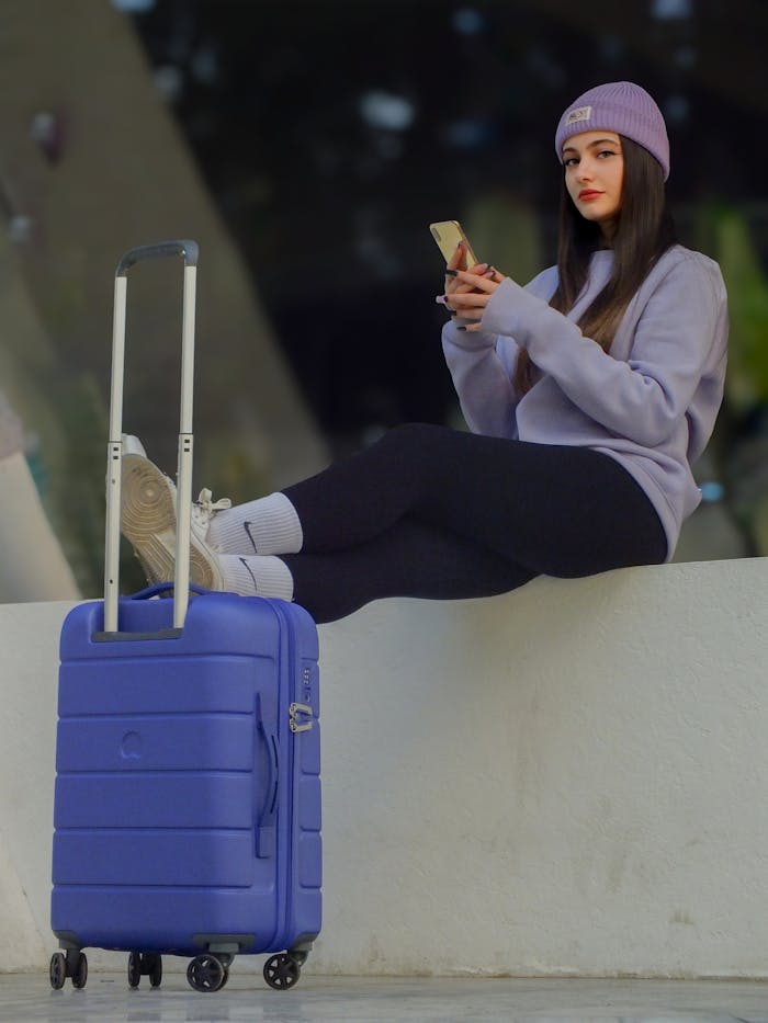 Casual young woman sitting with suitcase, engaging with her phone. Modern travel vibe.